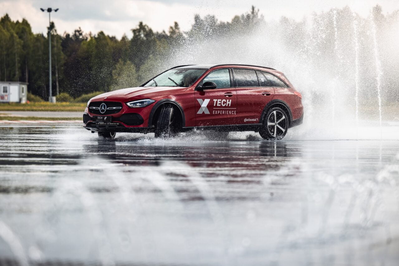 Czerwony samochód marki Mercedes przemierzający mokrą nawierzchnię z odbijającymi wodą i fontannami w tle.