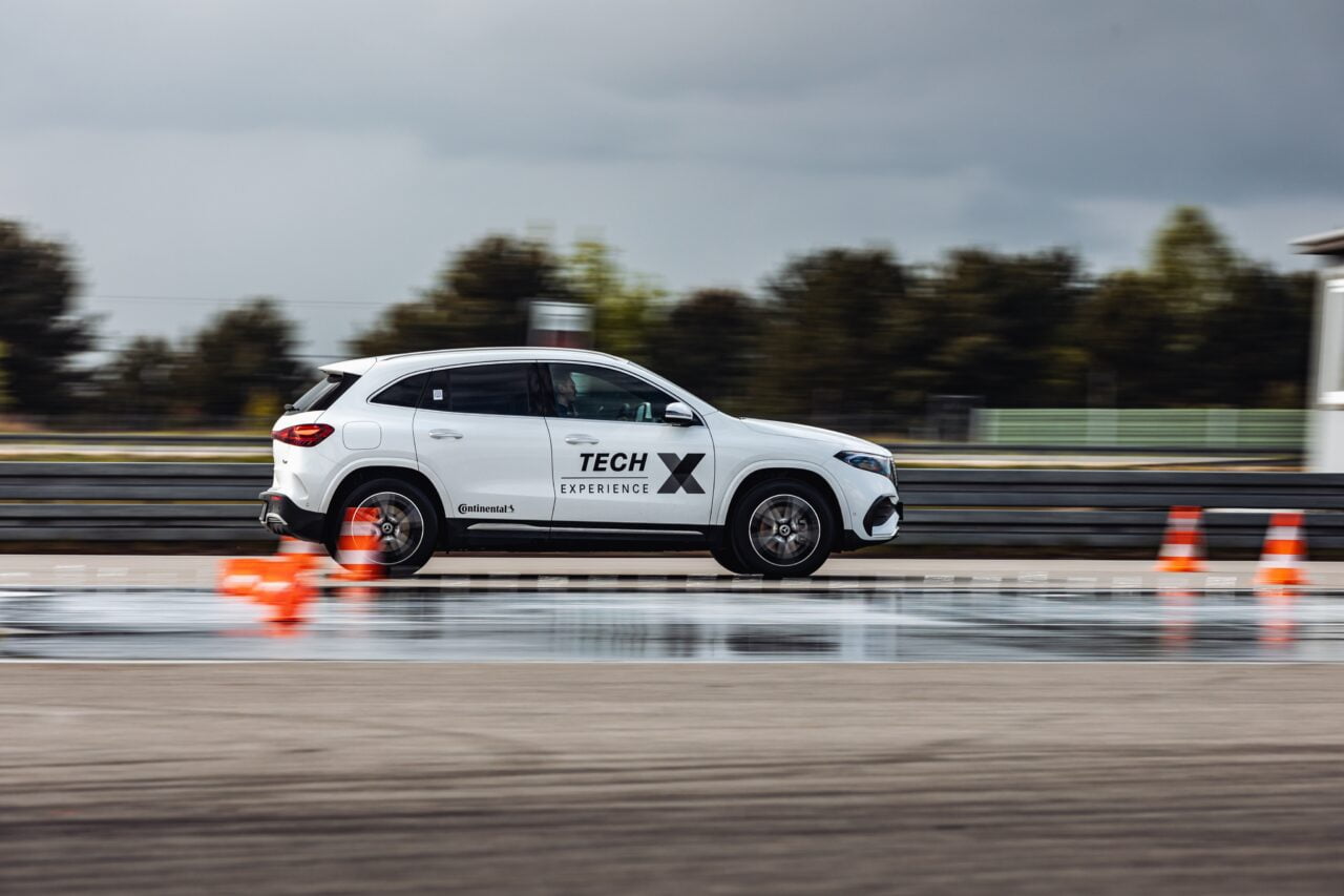 Biały SUV jadący z dużą prędkością na mokrym torze testowym, z napisami "Tech X Experience" na boku.