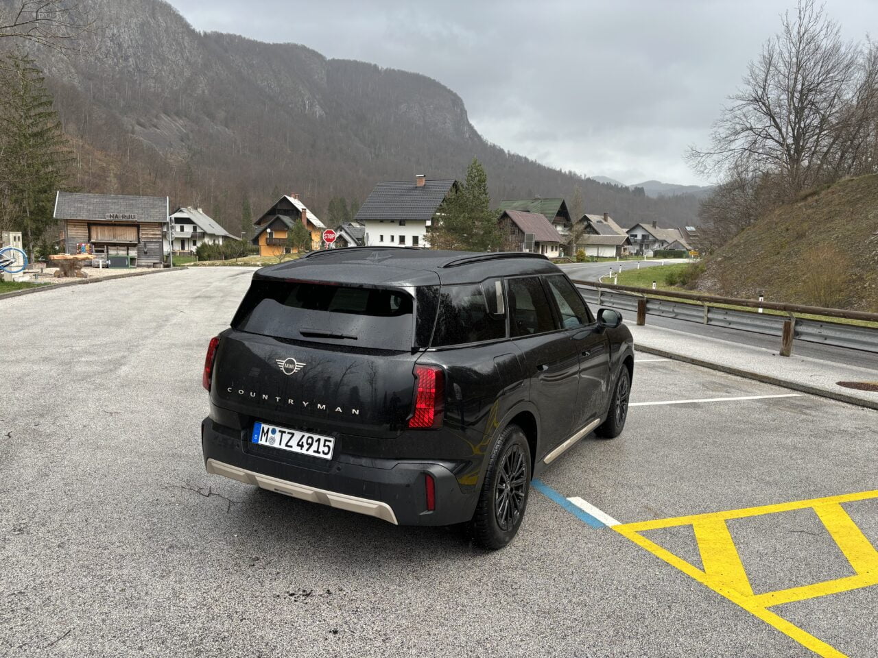 Czarny samochód marki Mini Countryman zaparkowany na poboczu drogi w górskiej scenerii, z widocznymi domami i szarym niebem w tle.