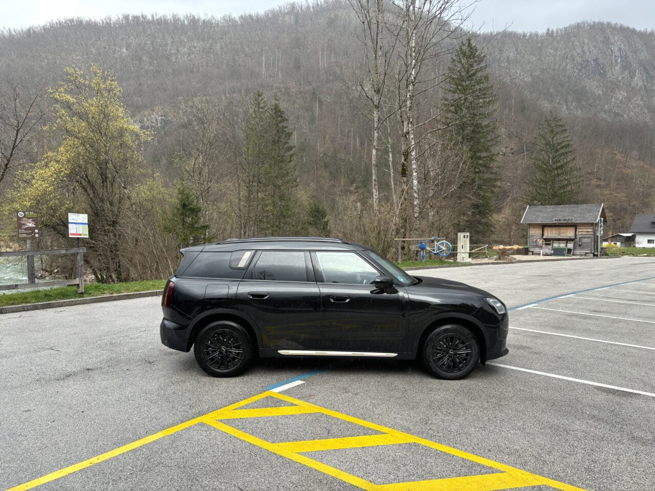 Czarny SUV zaparkowany na pustym parkingu z widokiem na góry i mały drewniany budynek obok rzeki.