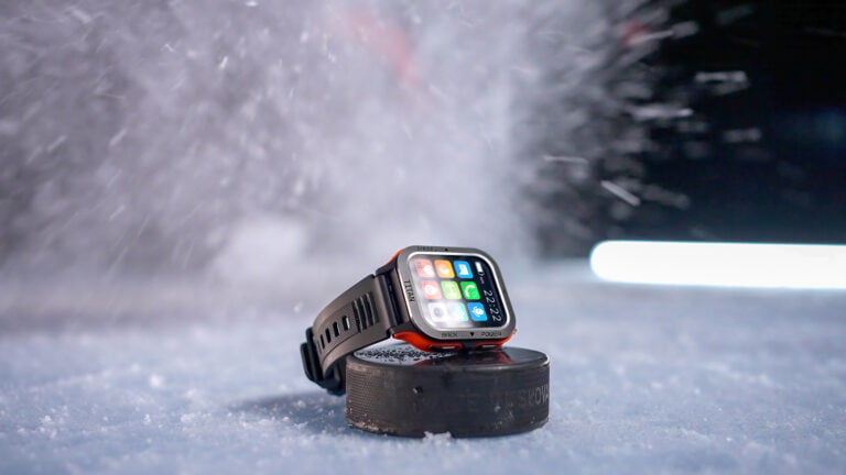 Smartwatch Maxcom FW67 Titan Pro umieszczony na czarnym krążku hokejowym na śnieżnej powierzchni z wymazanym ruchem śniegu w tle.