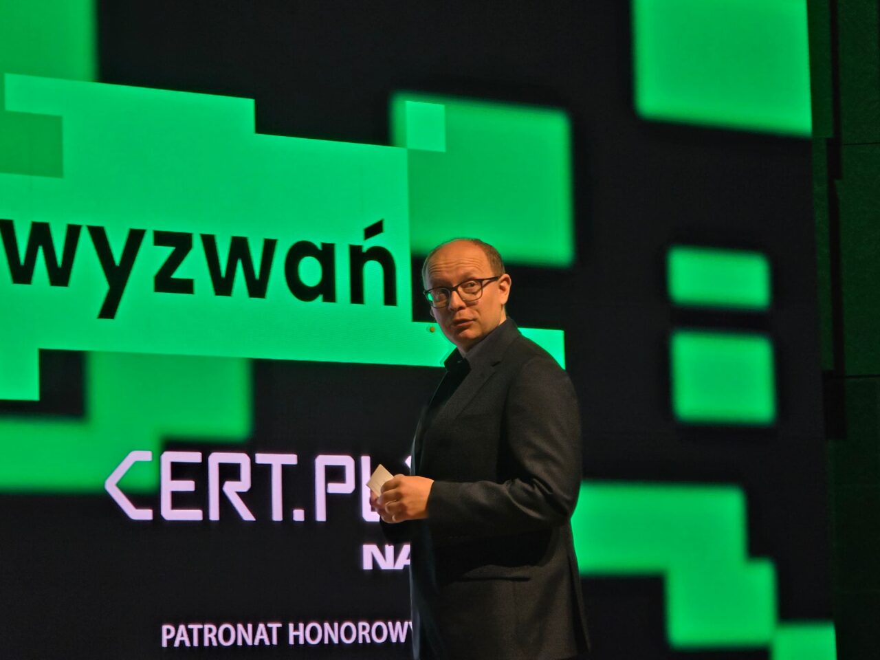 Mężczyzna w ciemnym garniturze stojący na scenie z mikrofonem w ręku na tle dużego ekranu wyświetlającego grafikę z tekstem "wyzwań" i logo CERT.PL.