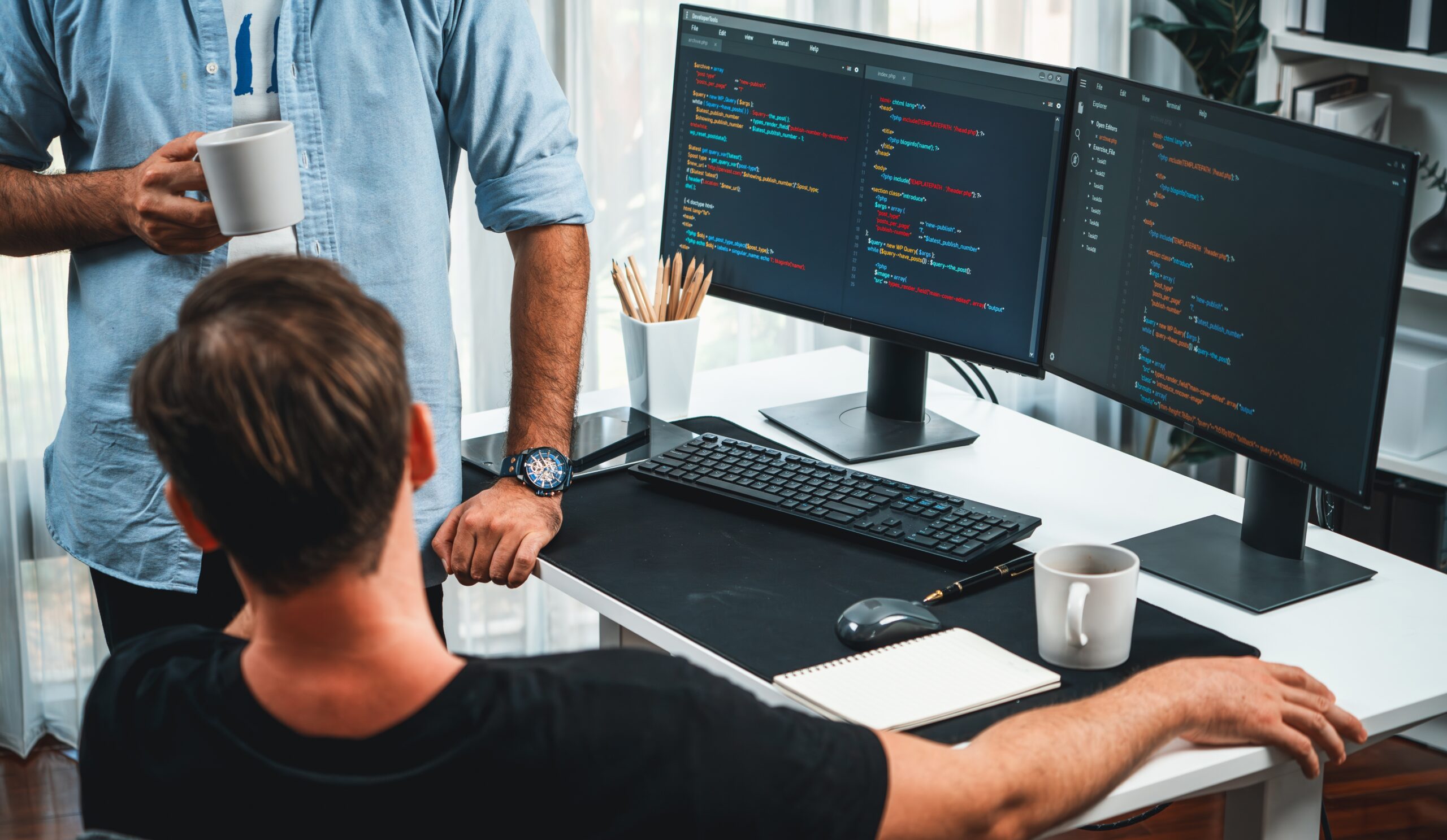 Dwóch mężczyzn pracuje przy biurku z dwoma monitorami wyświetlającymi kod programistyczny, jeden siedzi, drugi stoi z filiżanką kawy.