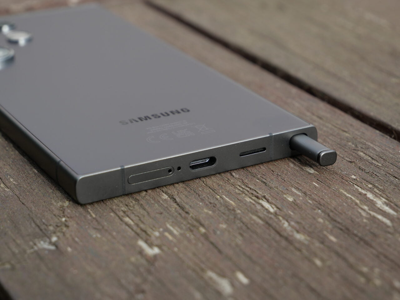 Szary smartfon marki Samsung leżący na drewnianej powierzchni z widocznym wyciągniętym rysikiem S Pen i portami na bocznym brzegu.