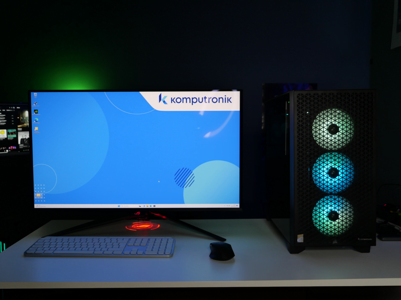Komputer stacjonarny z podświetlaną obudową i monitor na pulpicie z logo Komputronik, klawiatura i mysz na biurku, zielone podświetlenie w tle.