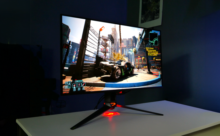 Monitor komputerowy z wyświetlonym obrazem gry Cyberpunk 2077, postawiony na biurku w oświetlonym wielokolorowym światłem pokoju.