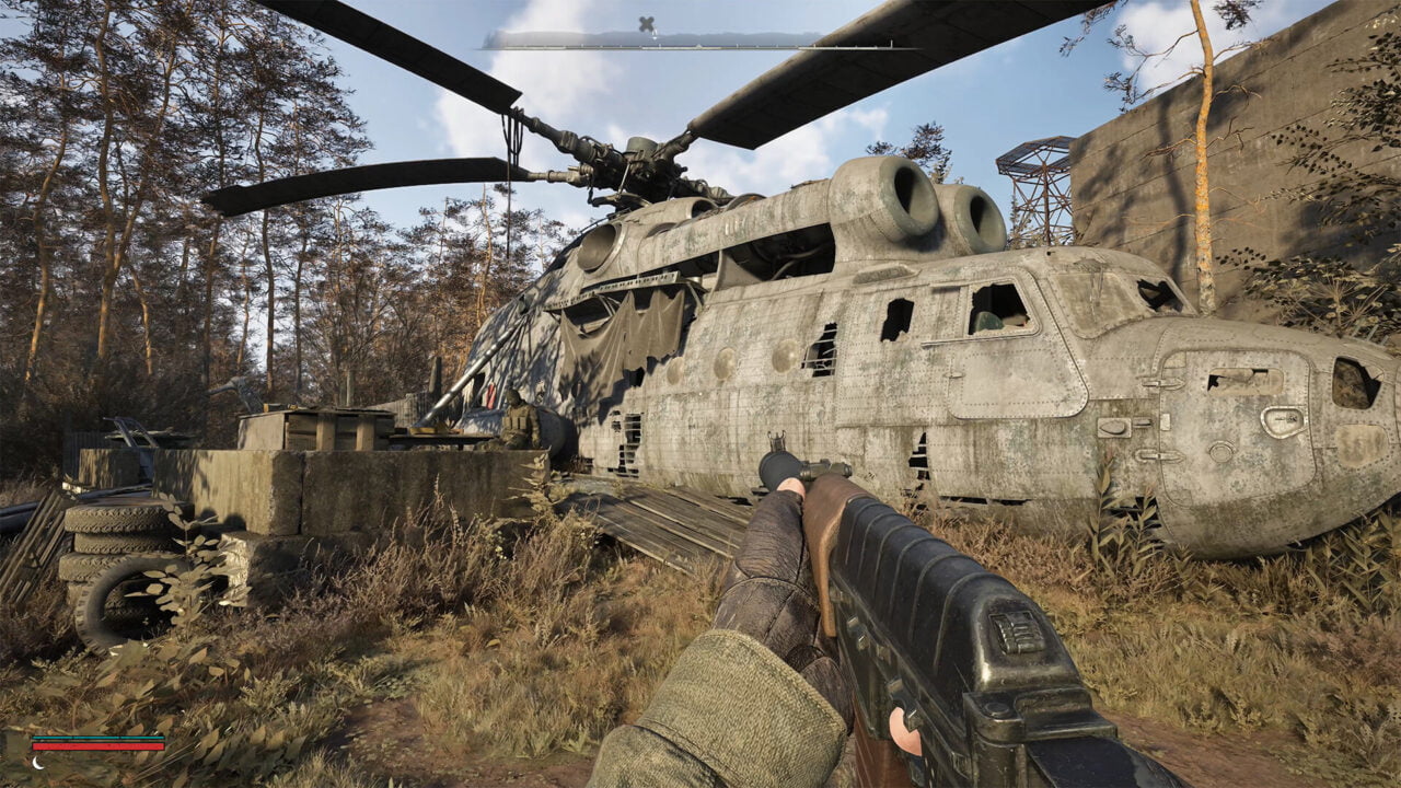 Kadr z gry STALKER 2. Perspektywa pierwszoosobowa postaci trzymającej broń, celującej w opuszczony duży śmigłowiec wojskowy w leśnym terenie.