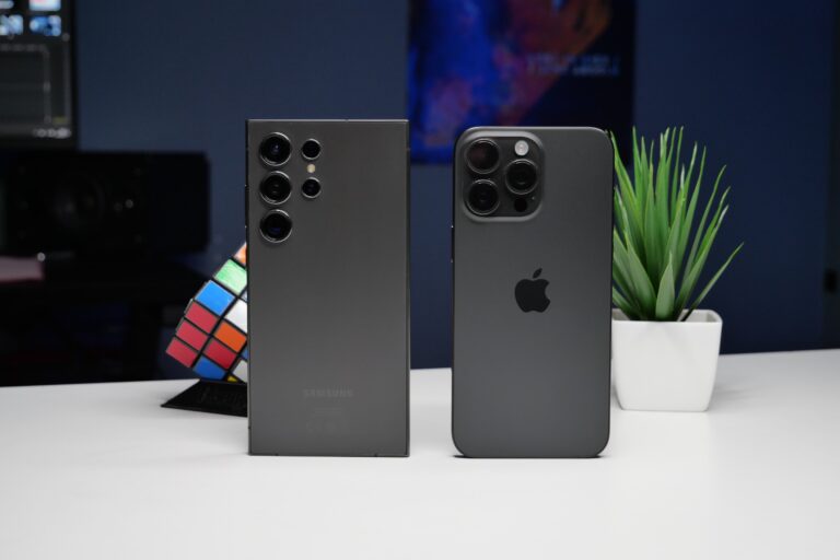 Dwa smartfony stoją tyłem na biurku; po lewej Samsung z potrójnym układem aparatów, po prawej iPhone z potrójną kamerą, w tle kolorowa kostka Rubika i roślina doniczkowa.
