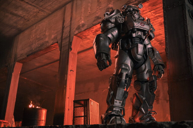 Kadr z serialu Fallout. Duży robot o futurystycznym wyglądzie stoi w półmroku przemysłowego wnętrza z czerwonym światłem w tle i palącą się beczką w pobliżu.