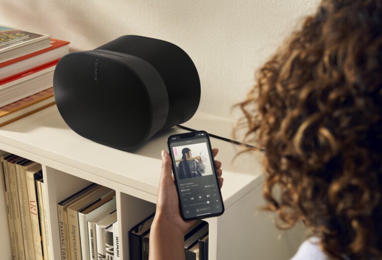 Kobieta z kręconymi włosami korzysta z aplikacji muzycznej na smartfonie w pobliżu czarnego głośnika Sonos umieszczonego na półce z książkami.