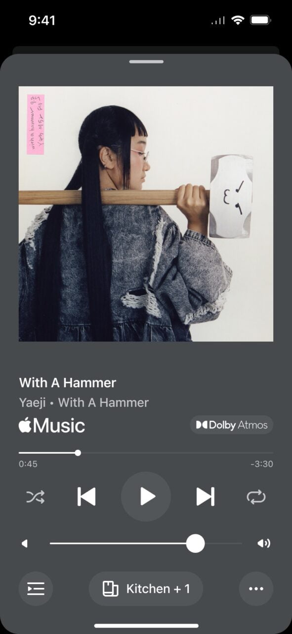 Nowa aplikacja Sonos. Kobieta w okularach i jeansowej kurtce trzyma pałkę, przykleja kartkę z symbolami na ścianę, ekran aplikacji muzycznej z utworem "With A Hammer" w tle.
