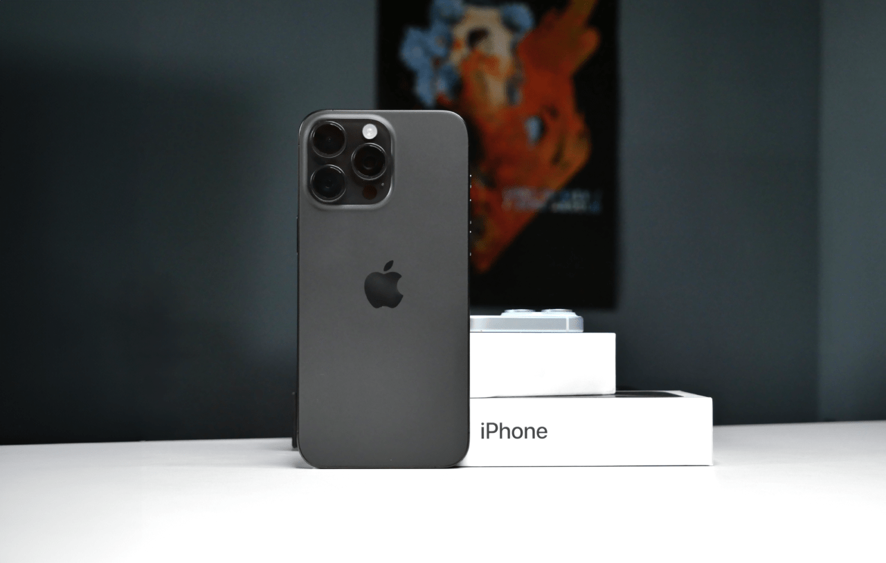 Czarny iPhone z potrójnym aparatem fotograficznym oparty o pudełko z napisem "iPhone", w tle rozmazane biurko i obraz.