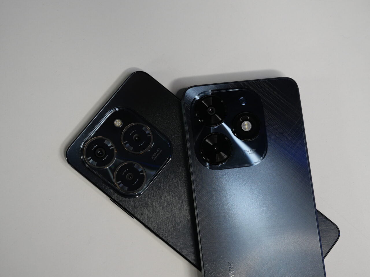 Dwa smartfony leżące na stole, jeden czarny i drugi niebieski, z widocznymi wieloma obiektywami aparatu na tylnym panelu.