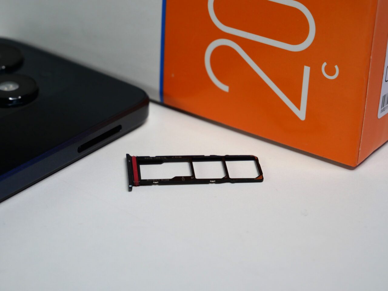 Część telefonu (tacka na kartę SIM) leży na stole obok czarnego smartfona i pomarańczowego pudełka z logo.