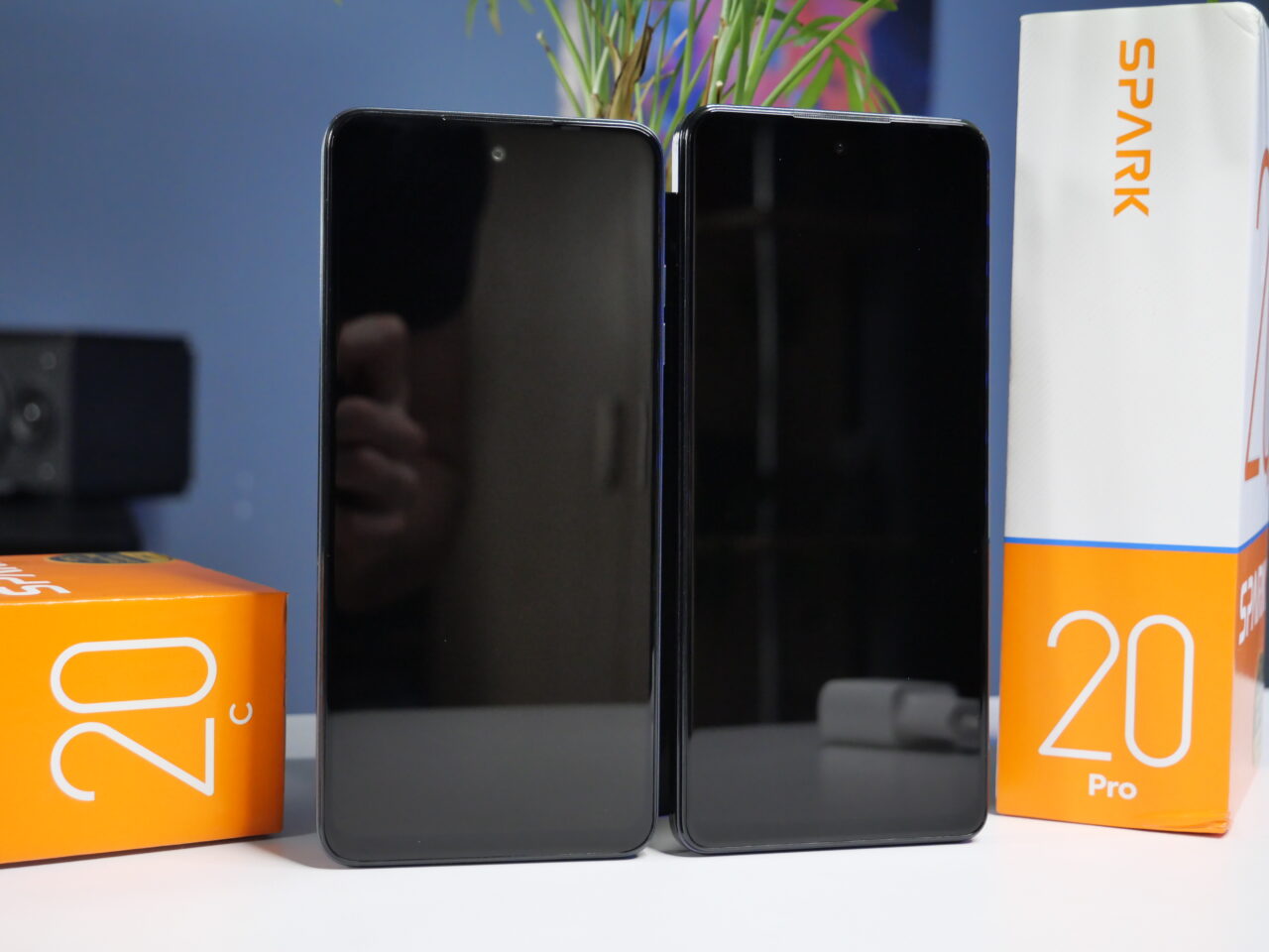 Dwa smartfony stoją pionowo obok siebie z opakowaniami Spark 20 i Spark 20 Pro, na tle niebieskiej ściany z małą rośliną.