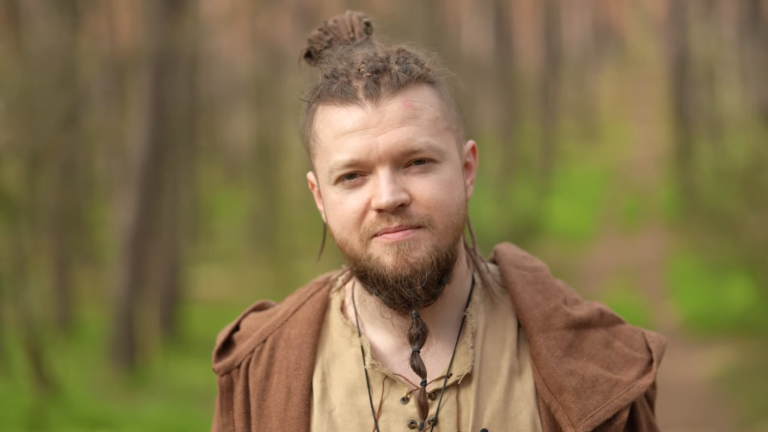Mężczyzna z brodą i wplecionymi włosami w stylu wikingów, ubrany w brązową tunikę, stoi w lesie.