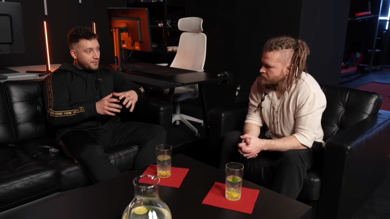 Dwóch mężczyzn siedzących naprzeciwko siebie w czarnym, nowoczesnym wnętrzu i prowadzących rozmowę, z napojami na stoliku przed nimi.