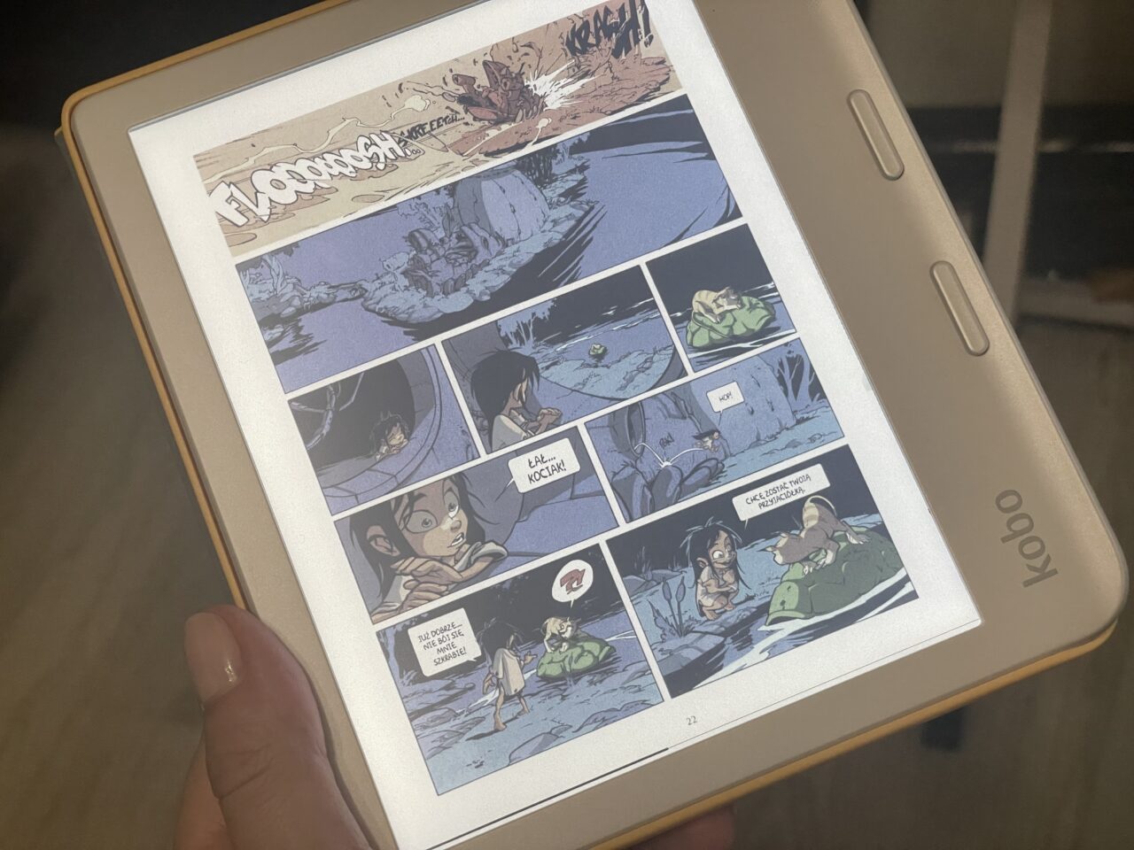 Ręka trzyma czytnik e-booków z wyświetlaną stroną komiksu zawierającą wiele kolorowych paneli z postaciami i tekstami.
