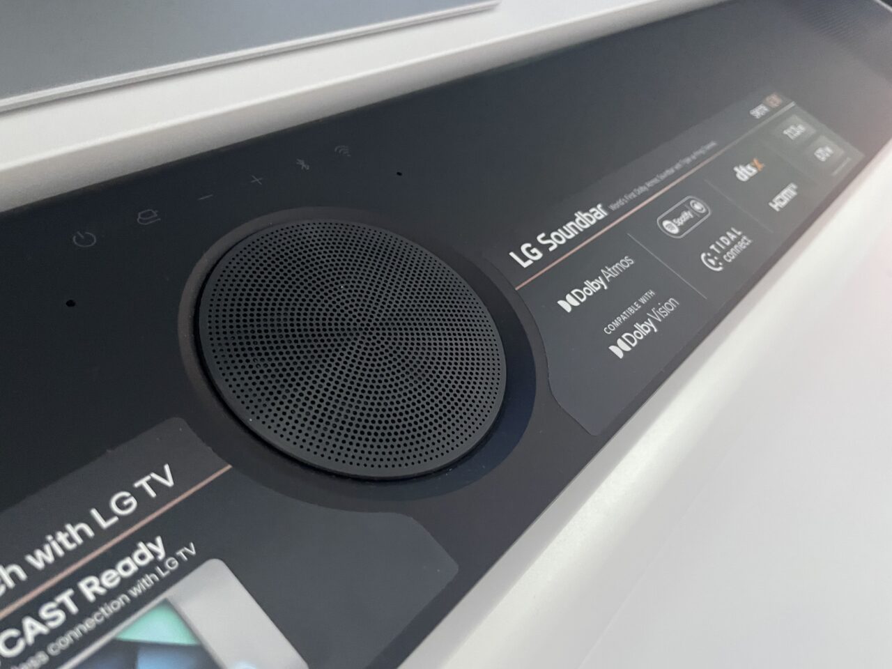 Zbliżenie na część górnej powierzchni czarnego soundbaru LG z logo i funkcjami Dolby Atmos, Dolby Vision oraz symbolami sterowania.