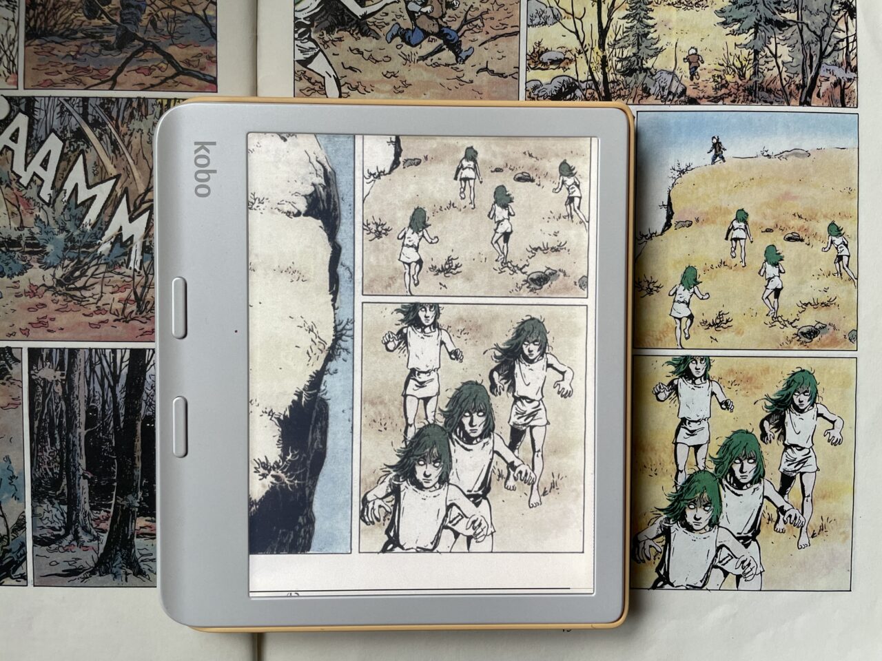 Czytnik e-booków Kobo wyświetlający stronę komiksu z przedstawiającymi bieganie postaciami na tle lasu i otwartej przestrzeni.