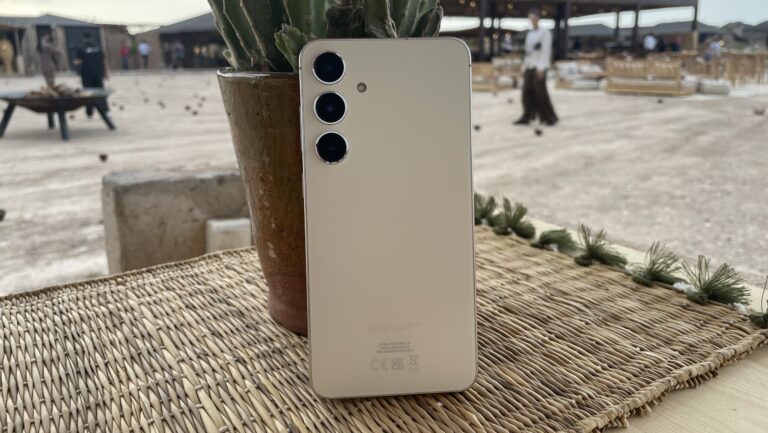 Biały smartfon z potrójnym aparatem ułożony na bambusowej matce obok doniczki z rośliną na drewnianym stole.