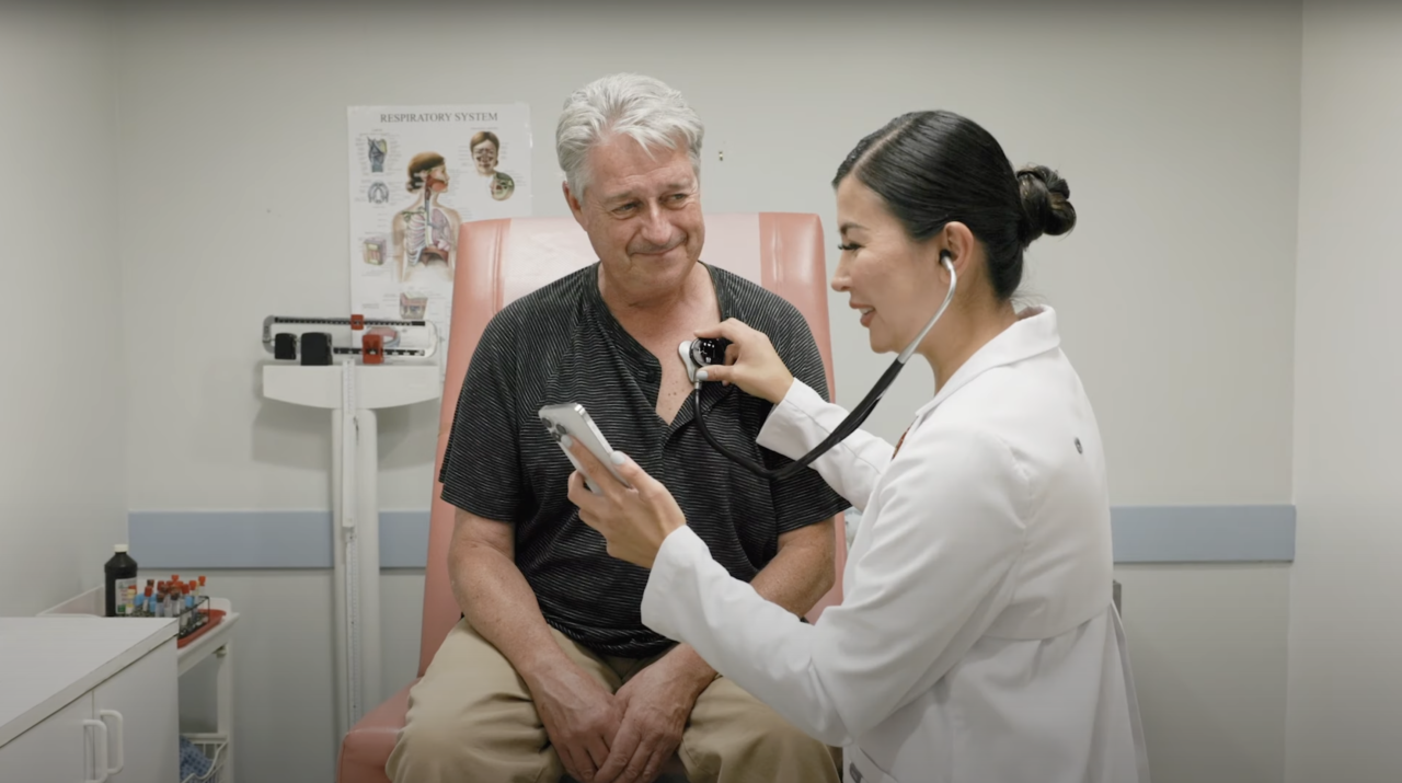 Starszy mężczyzna siedzący na fotelu lekarskim, podczas gdy lekarka bada go wykorzystując inteligentny stetoskop Eko Sensora i przegląda dane na telefonie w gabinecie lekarskim.