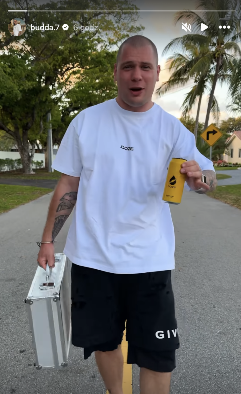 Mężczyzna trzymający metalową walizkę i żółtą puszeczkę napoju, stojący na ulicy z palmami w tle.