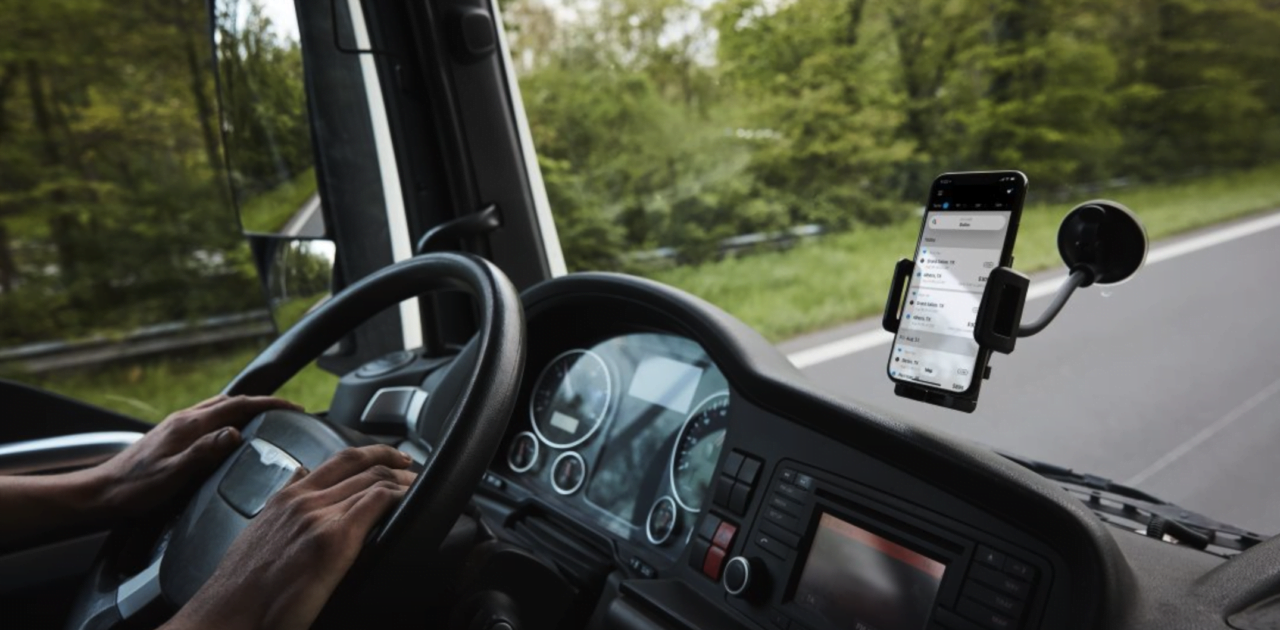 Kierowca trzyma kierownicę ciężarówki, a na przedniej szybie zamocowany jest telefon komórkowy w uchwycie z włączoną aplikacją nawigacyjną, widoczny z wnętrza kabiny.