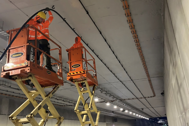 Pracownik w pomarańczowym odblaskowym ubraniu na podnośniku nożycowym wykonuje prace instalacyjne na suficie w tunelu.