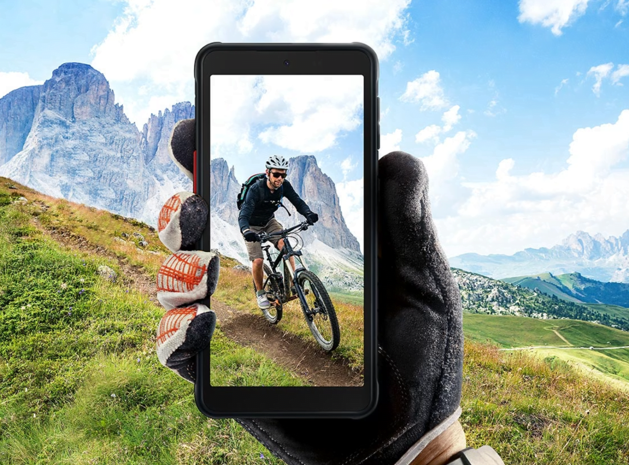 Ręka w rękawiczce trzymająca tablet pokazujący mężczyznę na rowerze górskim na tle górskiego krajobrazu.