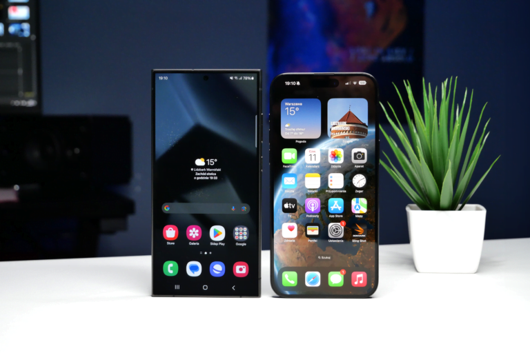 Dwa smartfony stojące na biurku, jeden z ekranem wyświetlającym aplikacje dla systemu Android, drugi z ekranem systemu iOS, obok doniczka z zieloną rośliną.
