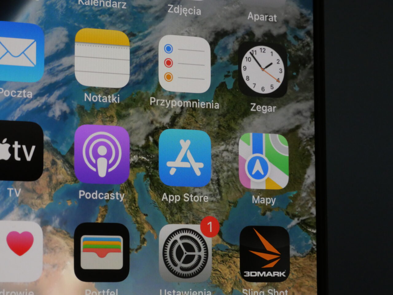 Zbliżenie na ekran urządzenia mobilnego z wyświetlonymi aplikacjami, takimi jak Kalendarz, Zdjęcia, Aparat, Notatki, Przypomnienia, Zegar, Podkasty, App Store, Mapy oraz inne.