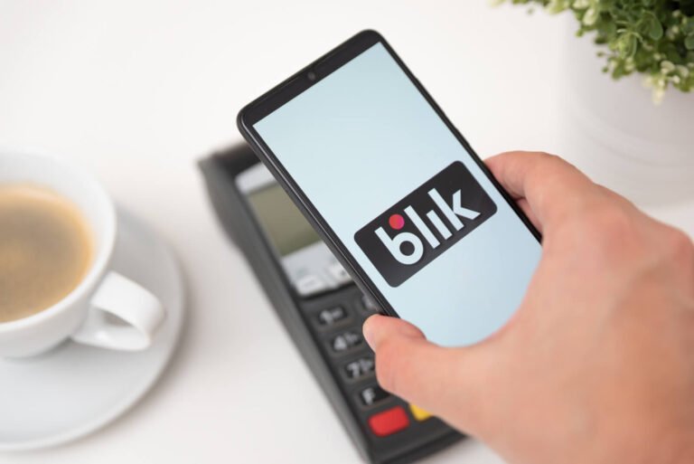 Osoba używa telefonu z wyświetlaczem pokazującym logo BLIK przy terminalu płatniczym, na biurku filiżanka kawy i doniczka z zieloną rośliną. BLIK nie działa