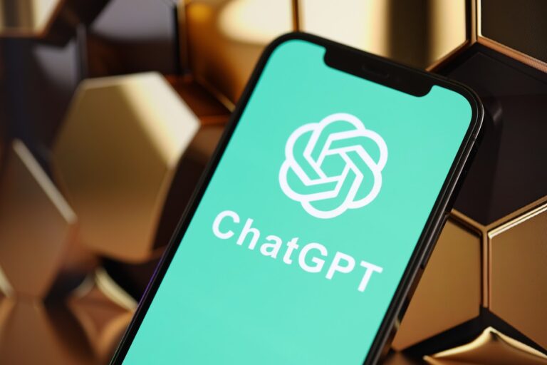 Smartfon z ekranem wyświetlającym logo ChatGPT, aplikacji stworzonej przez OpneAI, położony na tle metalicznych heksagonów.