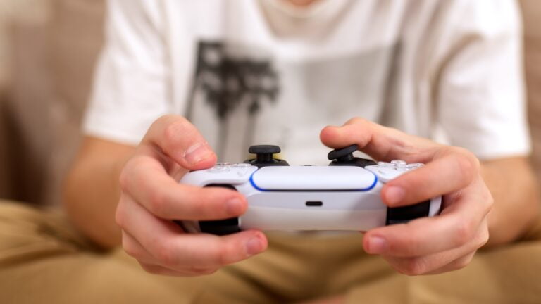 Osoba trzymająca biały kontroler do gier wideo, popularny w branży gier, w tle częściowo widoczna biała koszulka z nadrukiem