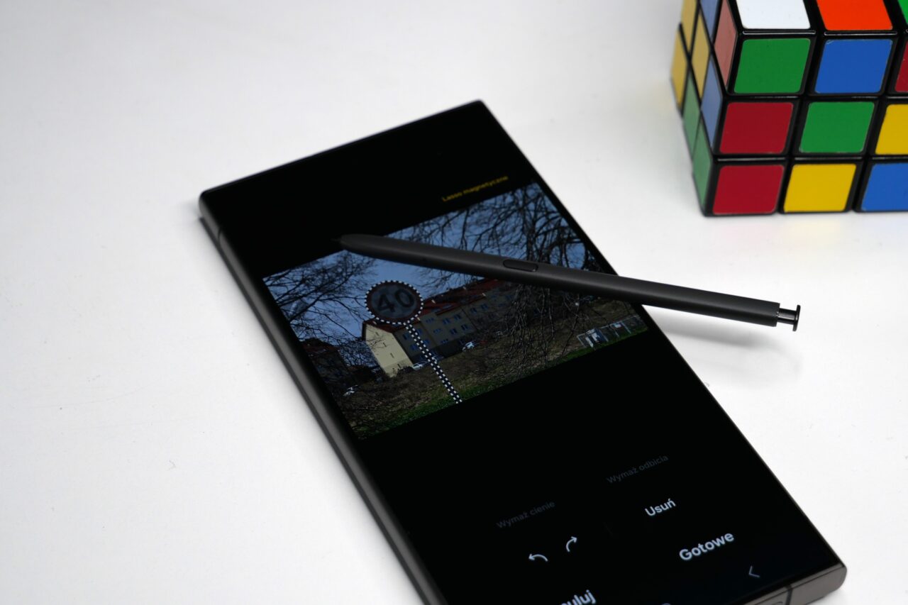 Czarny smartfon z rysikiem leżący na białym tle obok kostki Rubika, na ekranie zdjęcie wiosennej sceny miejskiej.