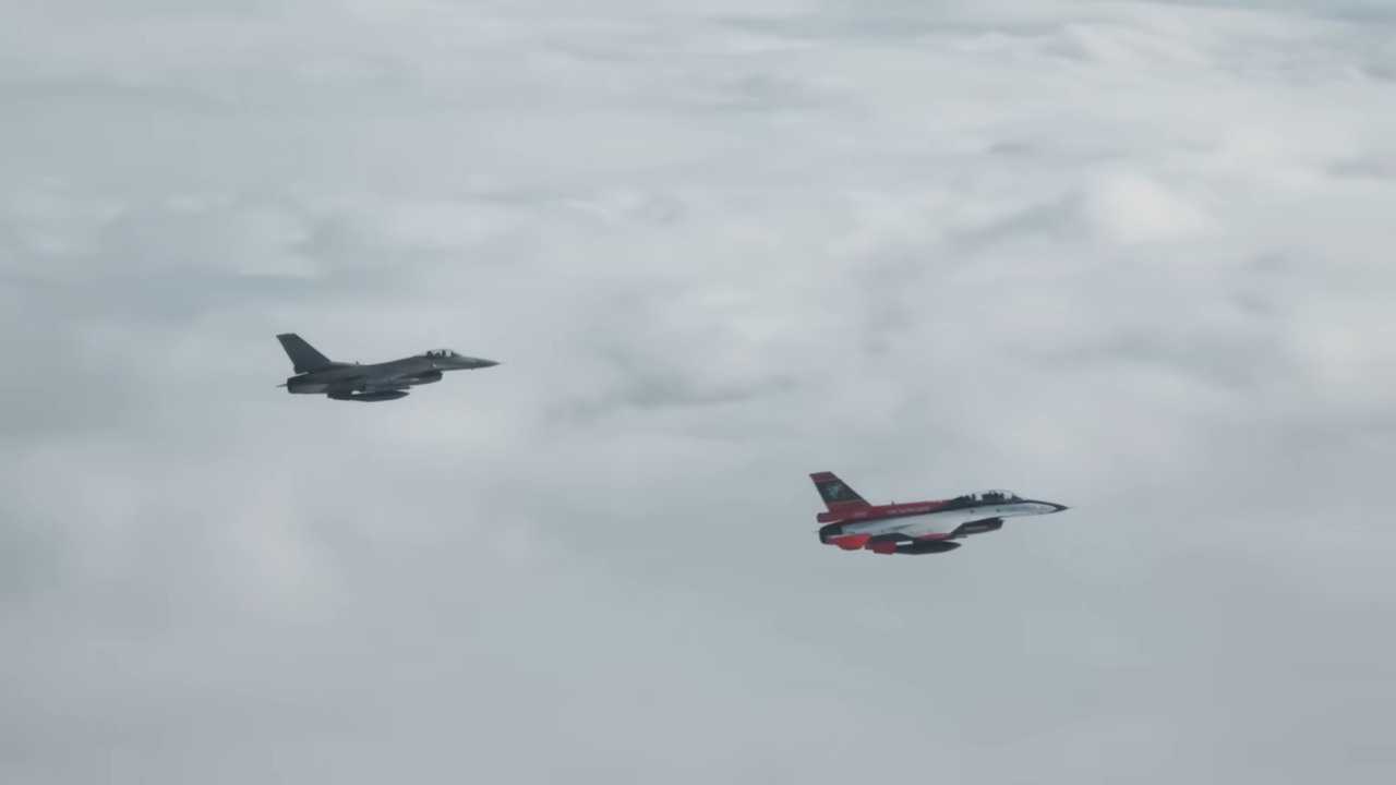 Dwa myśliwce F–16 lecące nad chmurami, jeden w barwach ciemnoszarych, drugi w czerwono-białych.