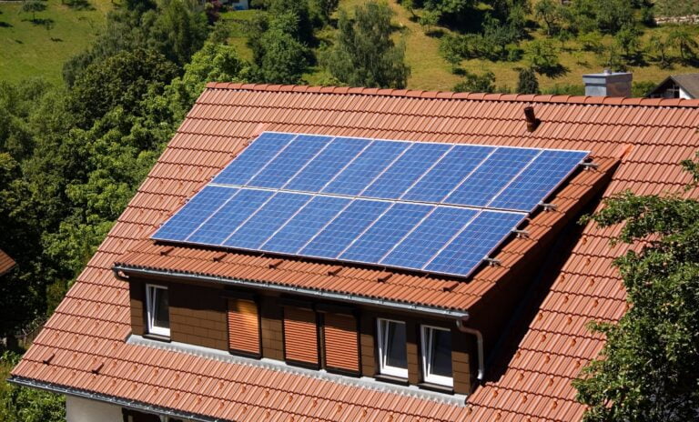 Panele słoneczne na dachu ceglanego domu otoczone zielenią.