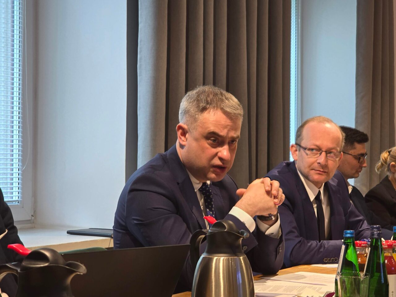 Minister Cyfryzacji Krzysztof Gawkowski podczas spotkania z mediami. Mężczyźni w garniturach siedzący przy stole konferencyjnym, jeden z nich patrzy na kamerę.