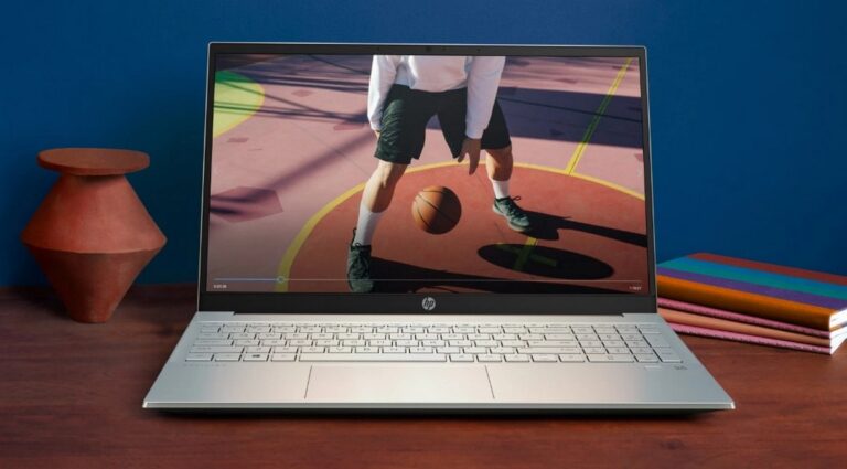Laptop HP na drewnianym biurku z wyświetlonym obrazem koszykarza odbijającego piłkę, obok czerwony wazon i kolorowe zeszyty.