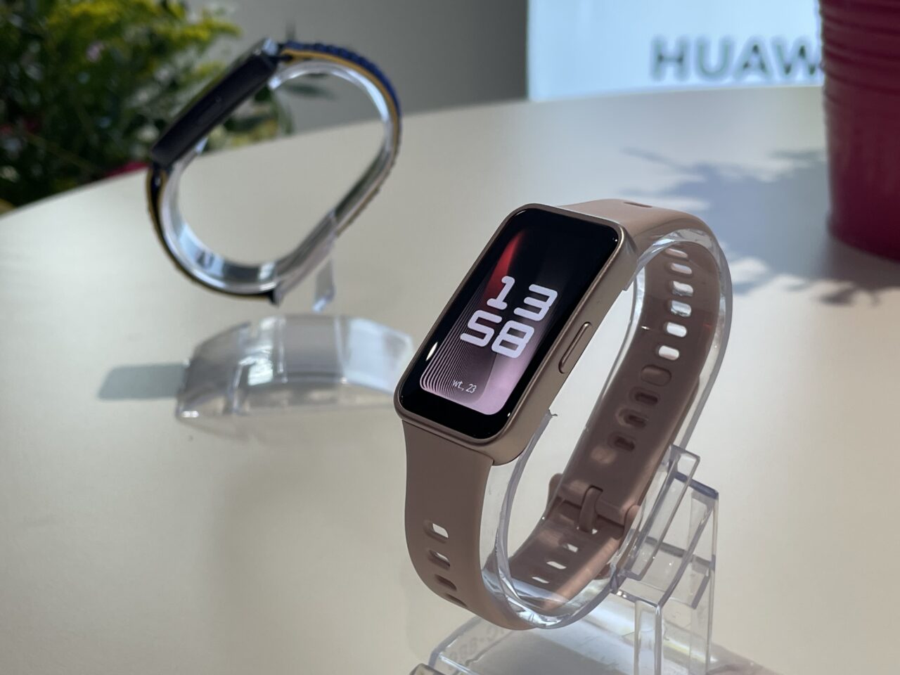 Smartwatch Huawei w różowym pasku na przezroczystym stojaku oraz drugi smartwatch z niebieskim paskiem w tle, na białym blacie przy naturalnym oświetleniu.