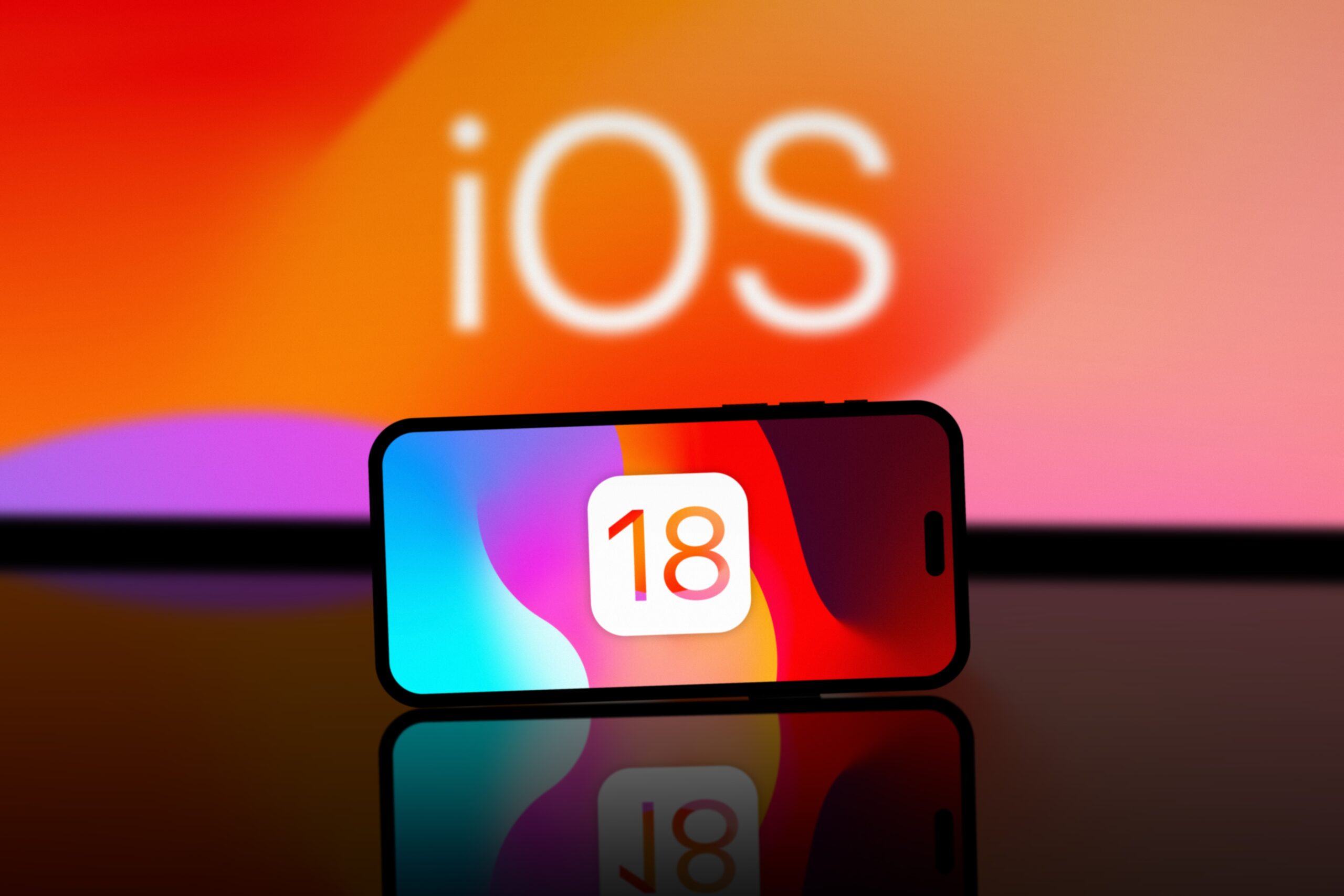 Telefon położony na refleksyjnej powierzchni z wyświetlanym logiem systemu iOS 18 na tle oświetlonym nasyconymi kolorami.