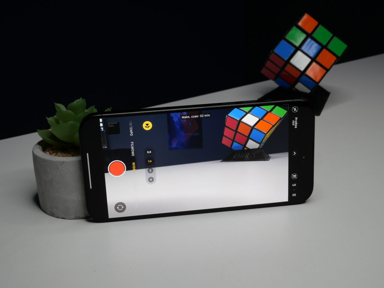Smartfon leżący na białym stole z włączoną aplikacją kamery wyświetlającą obraz kostki Rubika, obok doniczka z sukulentem.