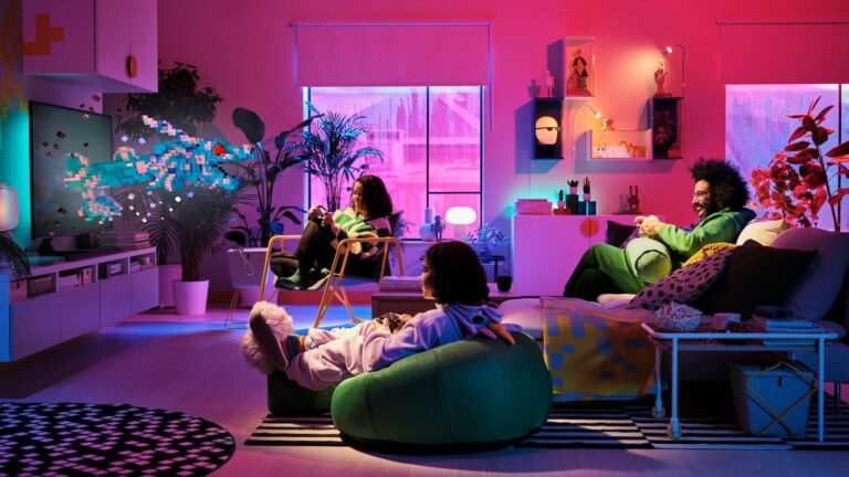 Nowoczesny pokój IKEA dzienny z neonowym różowym oświetleniem i trzema osobami grającymi w gry wideo. Interier zawiera półki z dekoracjami, dużą roślinę doniczkową, telewizor na ścianie i różnorodne, kolorowe meble. Atmosfera jest domowa i rozrywkowa.