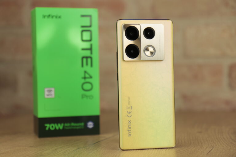 Testowany, złoty smartfon Infinix NOTE 40 Pro z potrójnym aparatem na tle pudełka z napisem Note 40 Pro i logo firmy na drewnianej powierzchni i rozmytym tle.