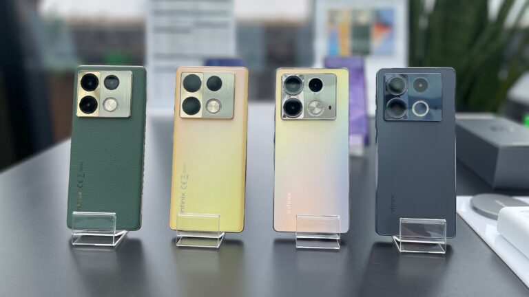 Cztery smartfony Infinix w różnych kolorach ustawione na przezroczystych podstawkach na stole.