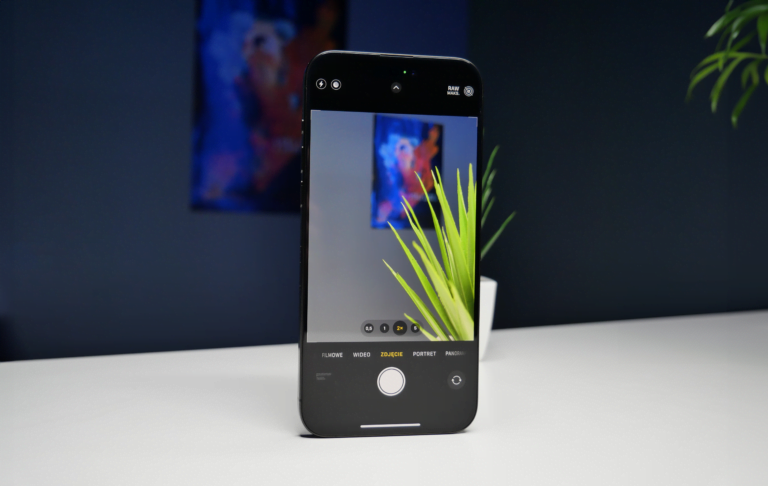 Smartfon umieszczony pionowo z otwartą aplikacją aparatu fotograficznego, który ma na celowniku doniczkę z zielonymi roślinami, w tle niebieskie tony i obraz.