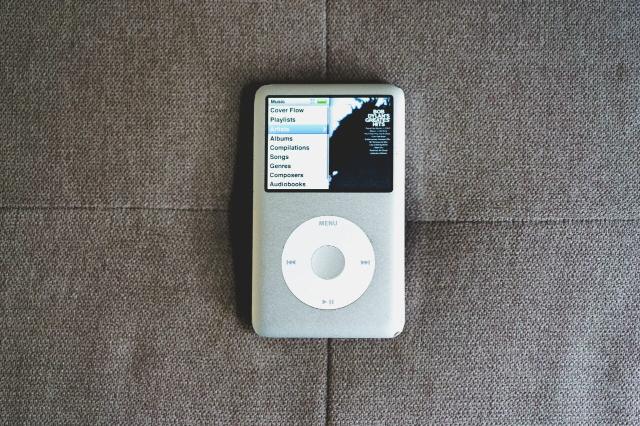 Srebrny odtwarzacz muzyki iPod Classic leżący na brązowej tkaninie, z wyświetlonym menu z muzyką.