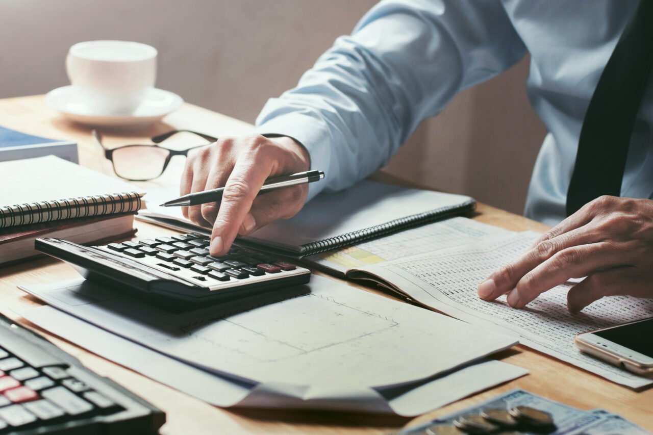 Mężczyzna w koszuli z krawatem, który pracuje przy biurku, używając kalkulatora i analizując dokumenty finansowe.