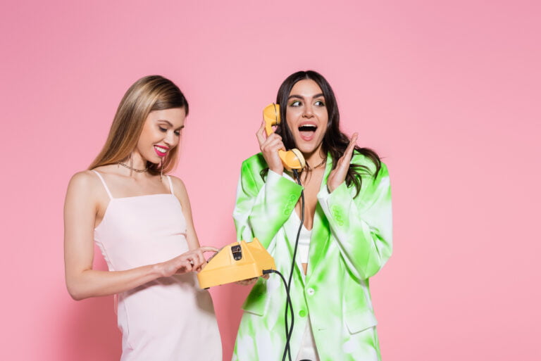 Dwie uśmiechnięte kobiety w modnych strojach z lat 80., jedna w białej sukience trzyma żółtą słuchawkę telefoniczną, a druga w zielonym garniturze mówi do niej, na różowym tle.